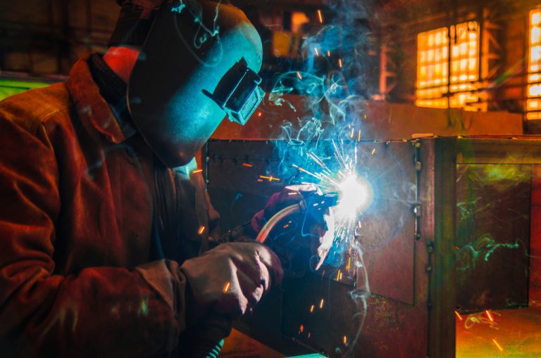 A welder wearing gloves and a mask uses MIG welding to attach a panel to furniture